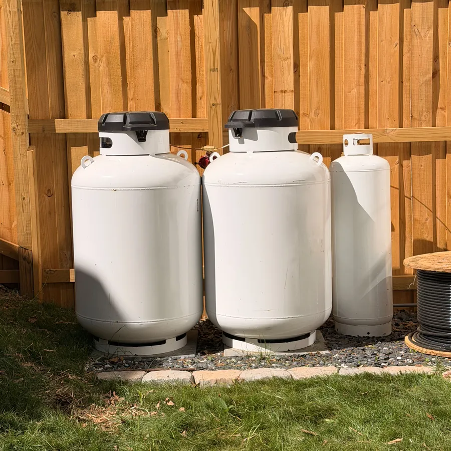 Propane tanks set on a gravel pad along a backyard fence to fuel a home standby generator